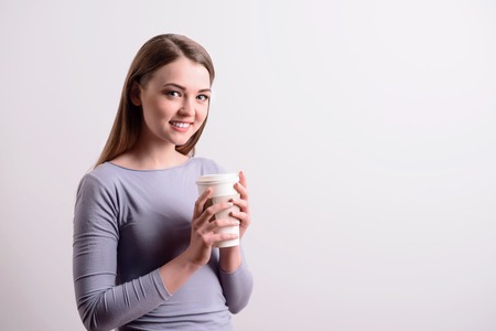 Cheerful beautiful positive woman smiling and drinking coffee while standing isolated on grey backgroundの写真素材