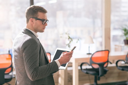 Pleasant serious concentrated man holding cell phone and using laptop while working in the office