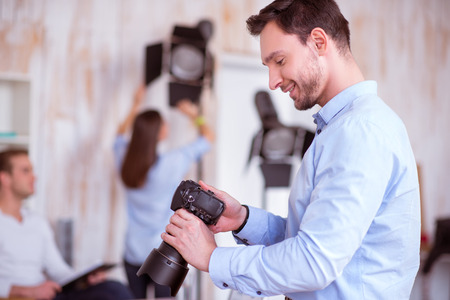 Pleasant smiling delighted handsome man holding photo camera and working in photo studio while...