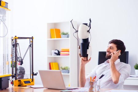 Pleasant positive handsome man holding model printed on 3d printer and sitting at the table while talking on cellphoneの写真素材