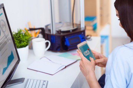 Pleasant beautiful woman sitting at the table and holding cellphone while 3d printer standing in the...