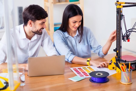 Pleasant smiling content colleagues sitting at the table and using 3d printer while expressing...