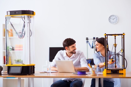 Cheerful delighted smiling colleagues sitting at the table and talking while using  3d printer