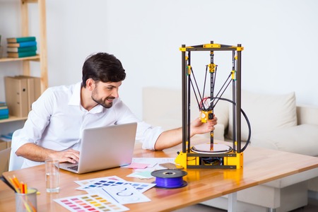 Cheerful delighted bearded smiling man sitting at the table and using 3d printer while working on...