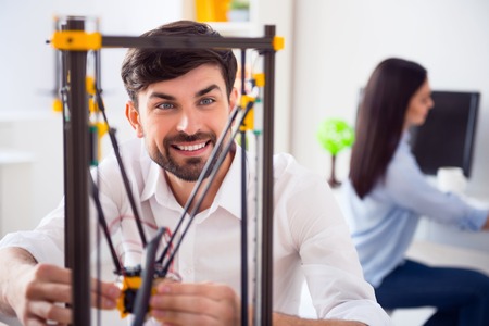 Cheerful delighted bearded smiling man sitting at the table and using 3d printer while his colleague...