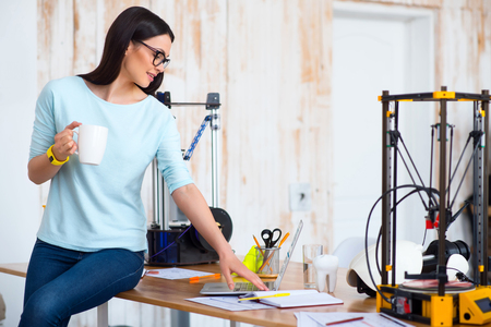 Cheerful beautiful woman sitting on the table and using laptop while d printer standing in the...