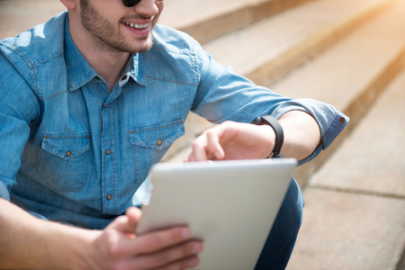 Gladness in mind. Cheerful content handsome smiling man looking at his wrist watch and holding tablet while sitting on the footstepsの写真素材