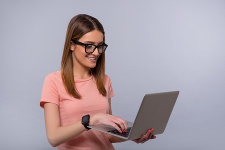 Involved in modern world. Positive delighted smiling woman using laptop and expressing gladness while standing isolated on grey backgroundの写真素材