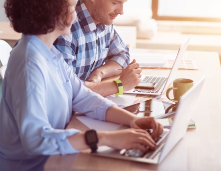 Work in concert. Pleasant delighted positive colleagues sitting at the table and using laptops while working in the officeの写真素材