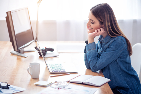Do everything with pleasure. Pleasant cheerful delighted smiling woman sitting at the table and using laptop while expressing positivityの写真素材