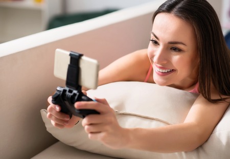 Playful mood. Cheerful delighted pleasant woman lying on the sofa and using game console while restingの写真素材