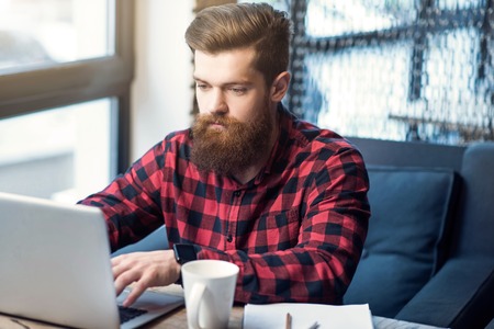 Keep modern pace. Pleasant delighted handsome  bearded man working on the laptop while sitting at the tableの写真素材