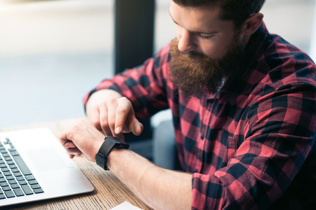 In touch with  the world. Pleasant bearded man sitting at the table and using smart watchの写真素材