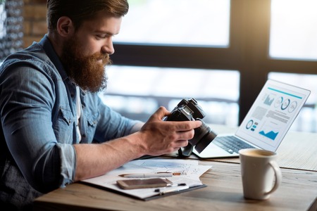 Charged with positivity. Pleasant content bearded man sitting at the table and holding photo camera while feeling gladの写真素材