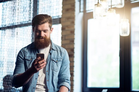 Feel the sound. Pleasant delighted bearded man standing in the office and holding cell phone while listening to musicの写真素材