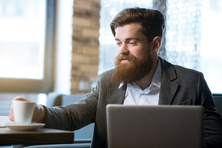 Have a nice day. Pleasant cheerful delighted bearded man drinking coffee while using laptopの写真素材