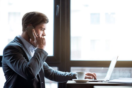 Involved in conversation. Pleasant bearded handsome man sitting at the table and working on the laptop while talking on cell phoneの写真素材