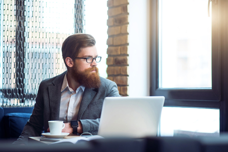 Serious attitude.   Pleasant concentrated bearded serious man sitting at the table looking aside while being involved in thinkingの写真素材