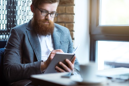 Plan your day. Pleasant glad bearded man sitting at the table and making notesの写真素材
