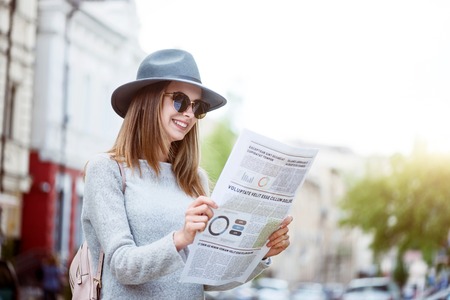 What is up. Smiling and merry young woman reading some articles in a newspaper while being in a bid modern cityの写真素材