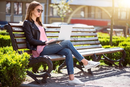 Beautiful day.  Positive and cheerful young woman using a laptop while sitting on a bench and  being in a parkの写真素材