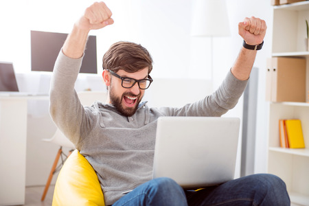 It is a victory. Lively young man looking at the screen while sitting on the armchair and raising his hands above the headの写真素材