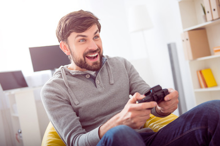Like a child. Joyful young bearded man sitting on the armchair and playing videogames