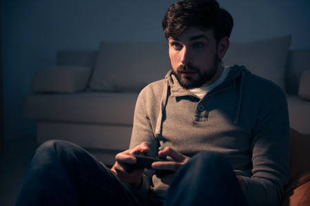 One more game. Young bearded man playing attentively while holding a videogame controller and...
