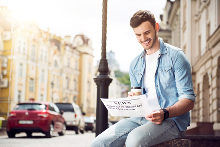 Daily press. Pleasant handsome delighted man sitting in the street and reading newspaper while drinking coffeeの写真素材