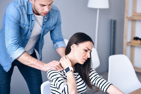Poor beautiful depressed woman sitting at the table and having pain in neck while the boyfriend making massage for herの写真素材