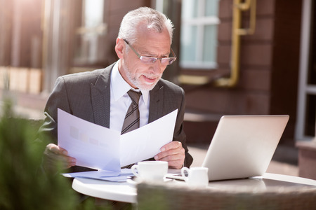 Pleasant work. Senior delighted businessman sitting at the table and using laptop while working with papersの写真素材