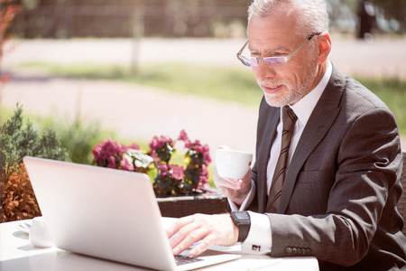 Refresh your mind. Pleasant content senior businessman sitting at the table and drinking coffee while using laptopの写真素材