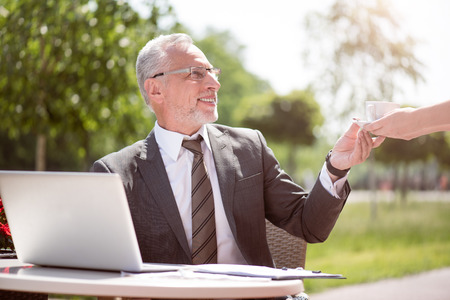best coffee for you. Positive delighted senior man sitting at the table and getting cup from the waiter while smilingの写真素材