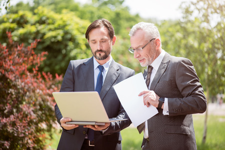 Pleasant work. Involved serious colleagues holding laptop and working outdoor while standing togetherの写真素材