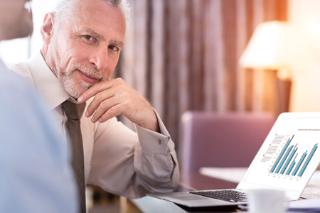 Confident glance. Pleasant delighted senior man sitting at the table and holding his hand near chin while using laptopの写真素材