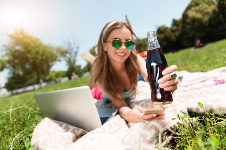 Favorite drink. Cheerful and smiling young woman holding cola bottle and her smart phone while lying on the grass in the parkの写真素材