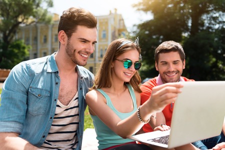 Stay up to date. Positive and delighted young woman and two young men using laptop during their studying while being in the park and sitting on the grassの写真素材