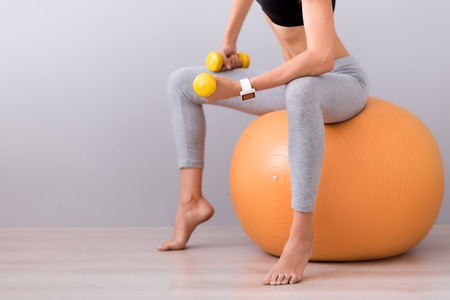 Stay in shape. Pleasant slim woman sitting on the rubber ball and using dumb bells while doing sport exercisesの写真素材