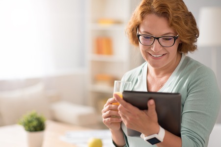True emotions. Cheerful delighted beautiful senior woman smiling and drinking juice while using tabletの写真素材