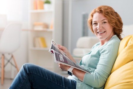 At home is the best. Joyful smiling senior woman sitting on the couch and reading magazine while restingの写真素材
