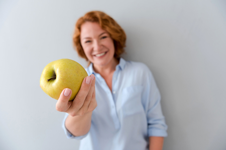 Here you are. Close up of apple in hand of pleasant cheerful senior woman holding it and smiling while standing isolated on grey backgroundの写真素材