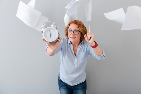 Take your time. Pleasant emotional senior woman holding alarm clock and pointing up while standing isolated on grey backgroundの写真素材