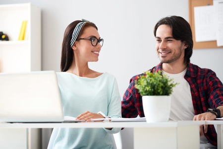 Cooperate with each other. Cheerful delighted smiling colleagues sitting at the table and using laptop while working togetherの写真素材