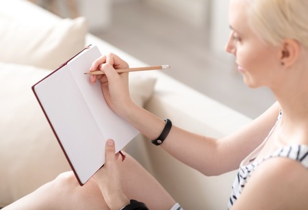 Lets note it. Amazing young woman looking down and taking notes in a notebook while sitting on the sofa at homeの写真素材