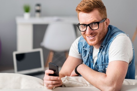 My helper. Cheerful handsome bearded man smiling and resting on the sofa while holding cell phone