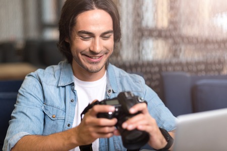 Real photographer. Cheerful delighted man sitting at the table and smiling while holding photo cameraの写真素材