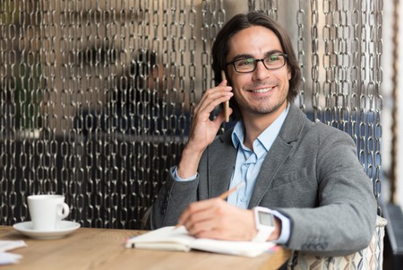 Glad to hear you. Cheerful content smiling man talking on cell phone and making notes while sitting in the cafeの写真素材