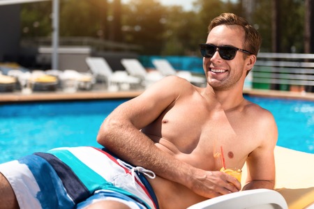 Open you mind. Overjoyed delighted man lying on the sun bed and drinking cocktail while resting near swimming poolの写真素材