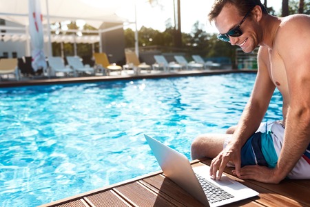 Modern user. Cheerful delighted handsome man sitting near swimming pool and resting while using laptopの写真素材