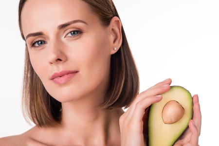 Exotic. Close up of cheerful young woman holding avocado and looking at a camera being on isolated...